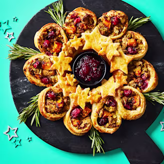 Puff pastry wreath made up of pastry cups and cut out stars assembled on a black serving board on a teal background.
