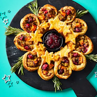 Puff pastry wreath made up of pastry cups and cut out stars assembled on a black serving board on a teal background.
