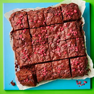 Chocolate brownie sliced into 16 squares with pink raspberries. On parchment paper and blue board with a green background.