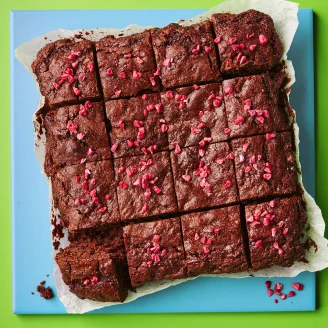 Chocolate brownie sliced into 16 squares with pink raspberries. On parchment paper and blue board with a green background.