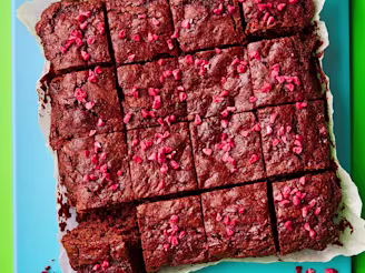 Chocolate brownie sliced into 16 squares with pink raspberries. On parchment paper and blue board with a green background.