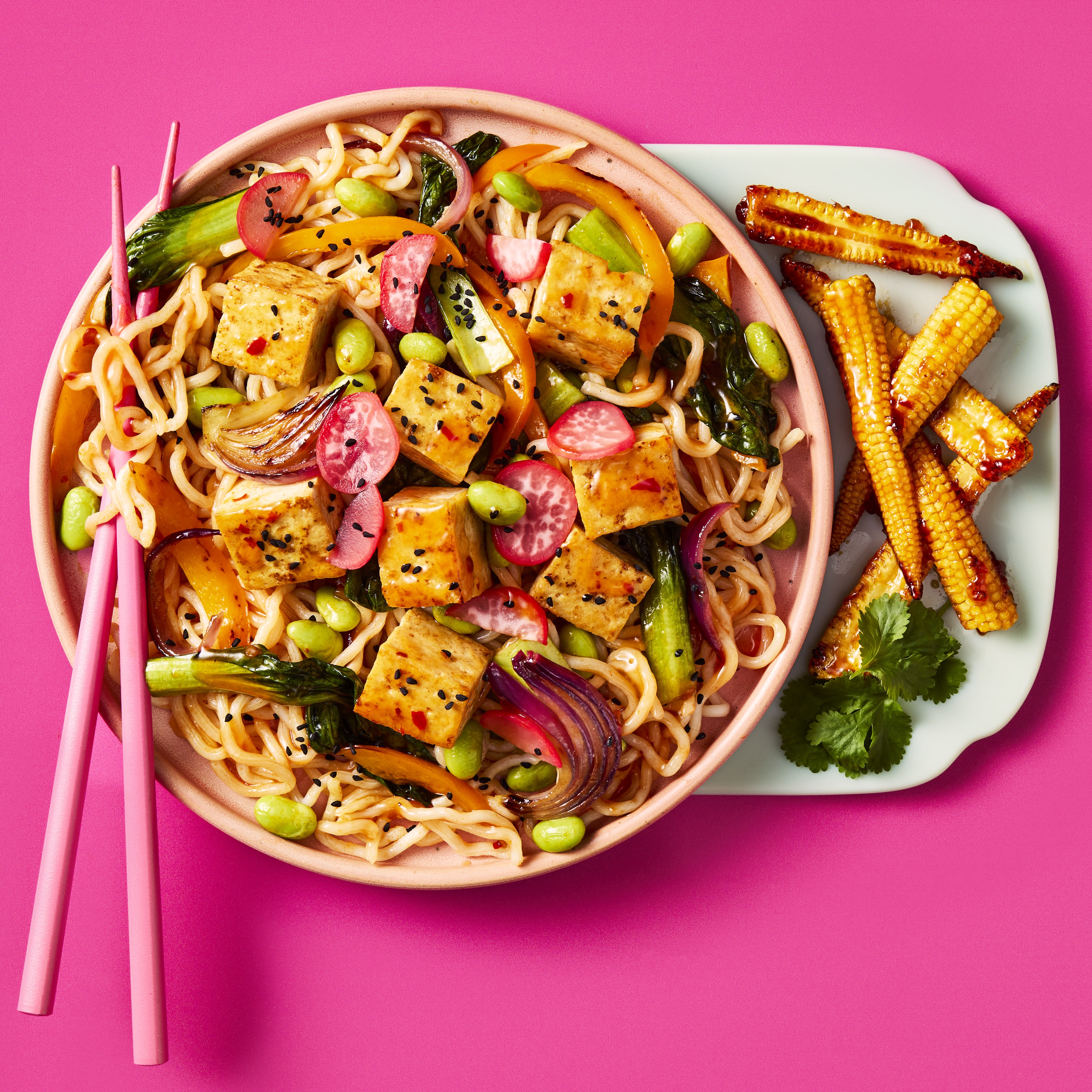 Cauldron Extra Firm Tofu cubed with noodles, radishes, pepper slices, edamame and baby corn with Sweet Chilli & Garlic Sauce in a light pink bowl on a bright pink background. Pink chopsticks rest on the left hand side of the bowl.