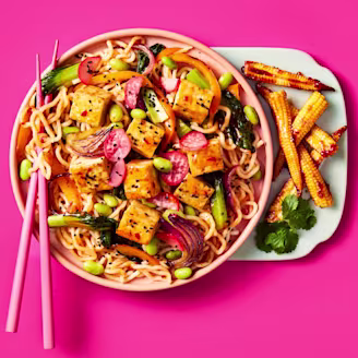 Cauldron Extra Firm Tofu cubed with noodles, radishes, pepper slices, edamame and baby corn with Sweet Chilli & Garlic Sauce in a light pink bowl on a bright pink background. Pink chopsticks rest on the left hand side of the bowl.