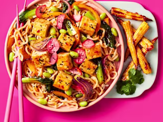 Cauldron Extra Firm Tofu cubed with noodles, radishes, pepper slices, edamame and baby corn with Sweet Chilli & Garlic Sauce in a light pink bowl on a bright pink background. Pink chopsticks rest on the left hand side of the bowl.