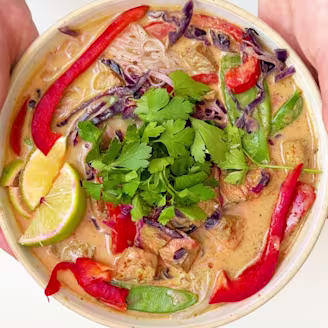 Orange tofu and coconut laksa curry in a bowl topped with sliced red pepper, lime wedges, fresh herbs and red cabbage.
