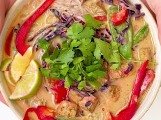 Orange tofu and coconut laksa curry in a bowl topped with sliced red pepper, lime wedges, fresh herbs and red cabbage.
