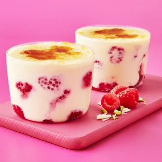 Two glass pots of crème brulee with raspberries on a pink board and background.