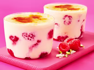 Two glass pots of crème brulee with raspberries on a pink board and background.