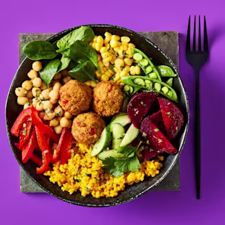 Three Cauldron Butternut & Red Chilli Bites in a black bowl on a slate placemat with bulgar wheat, sugar snap peas, sliced peppers, sweetcorn, cucumber, chickpeas, beetroot and coriander. A black fork is set to the right hand side on a purple background.