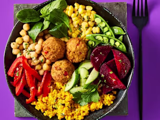 Three Cauldron Butternut & Red Chilli Bites in a black bowl on a slate placemat with bulgar wheat, sugar snap peas, sliced peppers, sweetcorn, cucumber, chickpeas, beetroot and coriander. A black fork is set to the right hand side on a purple background.