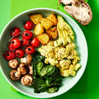 Cauldron Authentic Tofu breakfast bowl including potatoes, greens roasted mushrooms and tomatoes with a side of sourdough bread in a blue dish with a green background. 