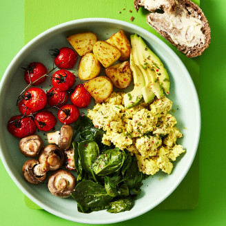 Cauldron Authentic Tofu breakfast bowl including potatoes, greens roasted mushrooms and tomatoes with a side of sourdough bread in a blue dish with a green background. 