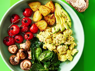 Cauldron Authentic Tofu breakfast bowl including potatoes, greens roasted mushrooms and tomatoes with a side of sourdough bread in a blue dish with a green background.