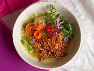 Brothy rice with grated tofu, sliced chilli, curled carrots and red onion in a white dish on a cream and pink background.