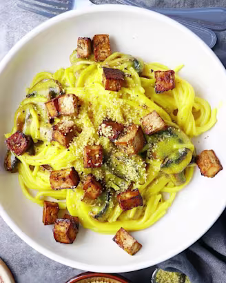Yellow carbonara spaghetti pasta with tofu in deep white bowls, small dish with parmesan cheese to side.