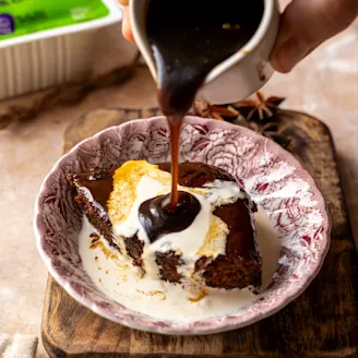 Sauce being poured on a vegan sticky toffee pudding made with tofu in a red ceramic bowl.
