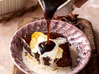 Sauce being poured on a vegan sticky toffee pudding made with tofu in a red ceramic bowl.
