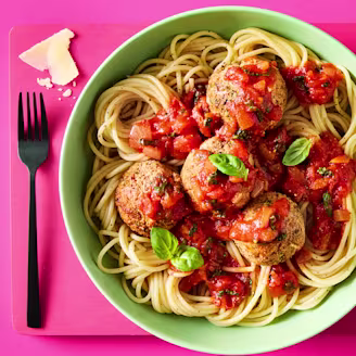 Tofu Meatballs & Pasta with parmesan and basil leaves in a green bowl on a pink background.