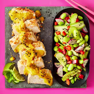 Sliced tofu topped with Katsu curry served alongside a salad including edamame, cucumber, radish and peppers, presented on a grey slab with a pink background.