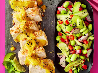 Sliced tofu topped with Katsu curry served alongside a salad including edamame, cucumber, radish and peppers, presented on a grey slab with a pink background.
