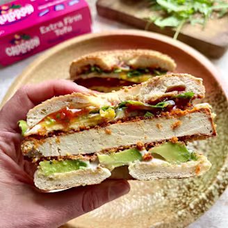 Hand holding bagel, with crispy tofu, avocado, pink Cauldron tofu packaging in background, greens on wooden chopping board.