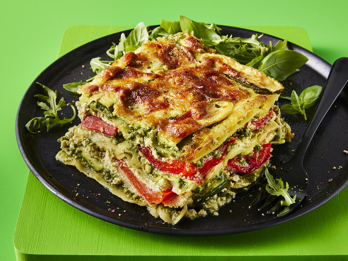 A slice of lasagne with the layers visible to showcase the fillings with a side of rocket served on a dark plate with a green background 
