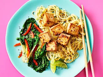 Cauldron tofu with noodles a side of pak choi and red peppers with sliced limes. Served on a blue plate and chopsticks on the side.