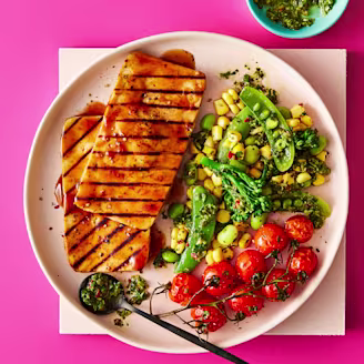 Two Tofu Steaks served alongside vegetables including green peans and tomatoes served on a white plate with a pink background 