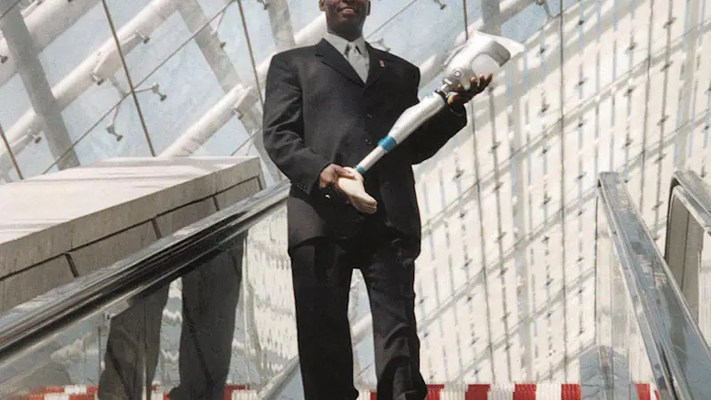 Curtis Grimsley steht mit Anzug auf einer Rolltreppe und hält eine Beinprothese in seinen Händen.