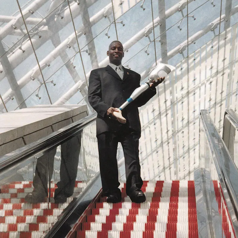 Curtis Grimsley steht mit Anzug auf einer Rolltreppe und hält eine Beinprothese in seinen Händen.