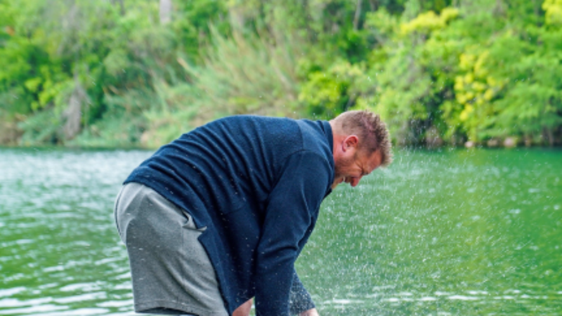 An Ottobock prosthetic leg user enjoys time outdoors by a lake with his dog. He bends down on a rock to pet the wet dog, smiling in a natural, playful moment.