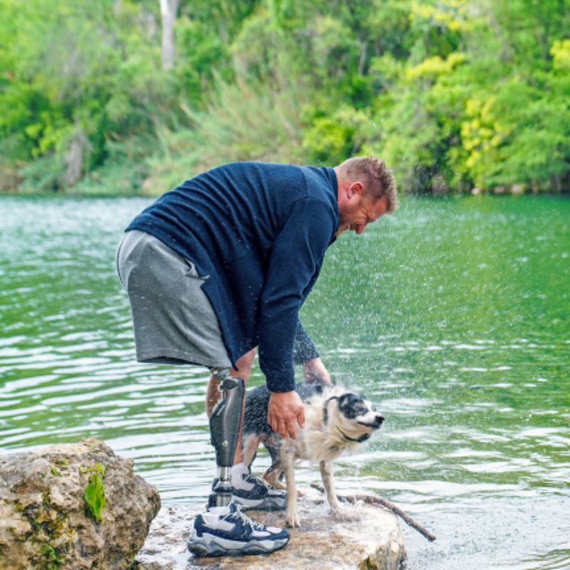 An Ottobock prosthetic leg user enjoys time outdoors by a lake with his dog. He bends down on a rock to pet the wet dog, smiling in a natural, playful moment.