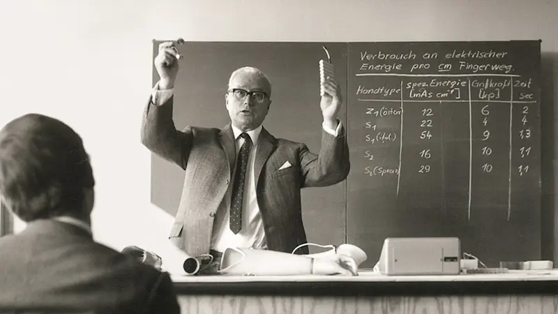 Dr. Max Näder steht an einer Tafel und hält mit gehobenen Händen einen Vortrag zu Myoelektrik.