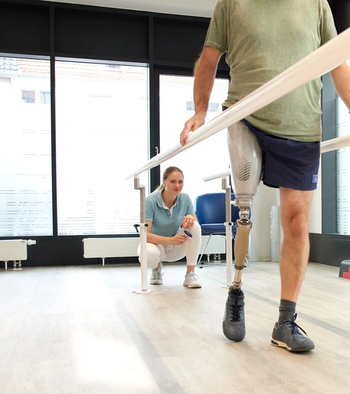 Man with above knee leg prosthesis walking between parallel bars during gait training, physiotherapist supervising in a rehabilitation clinic