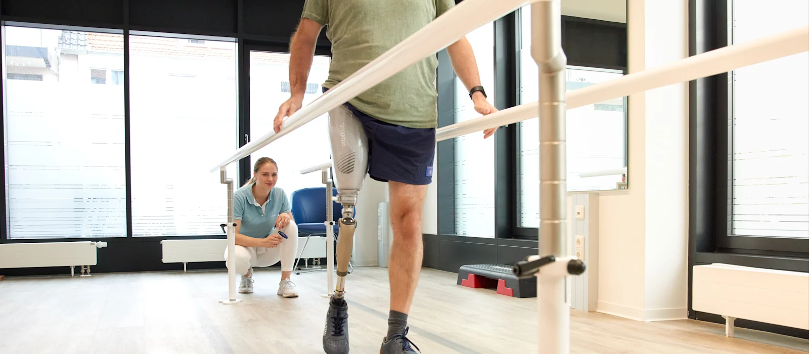 Man with above knee leg prosthesis walking between parallel bars during gait training, physiotherapist supervising in a rehabilitation clinic