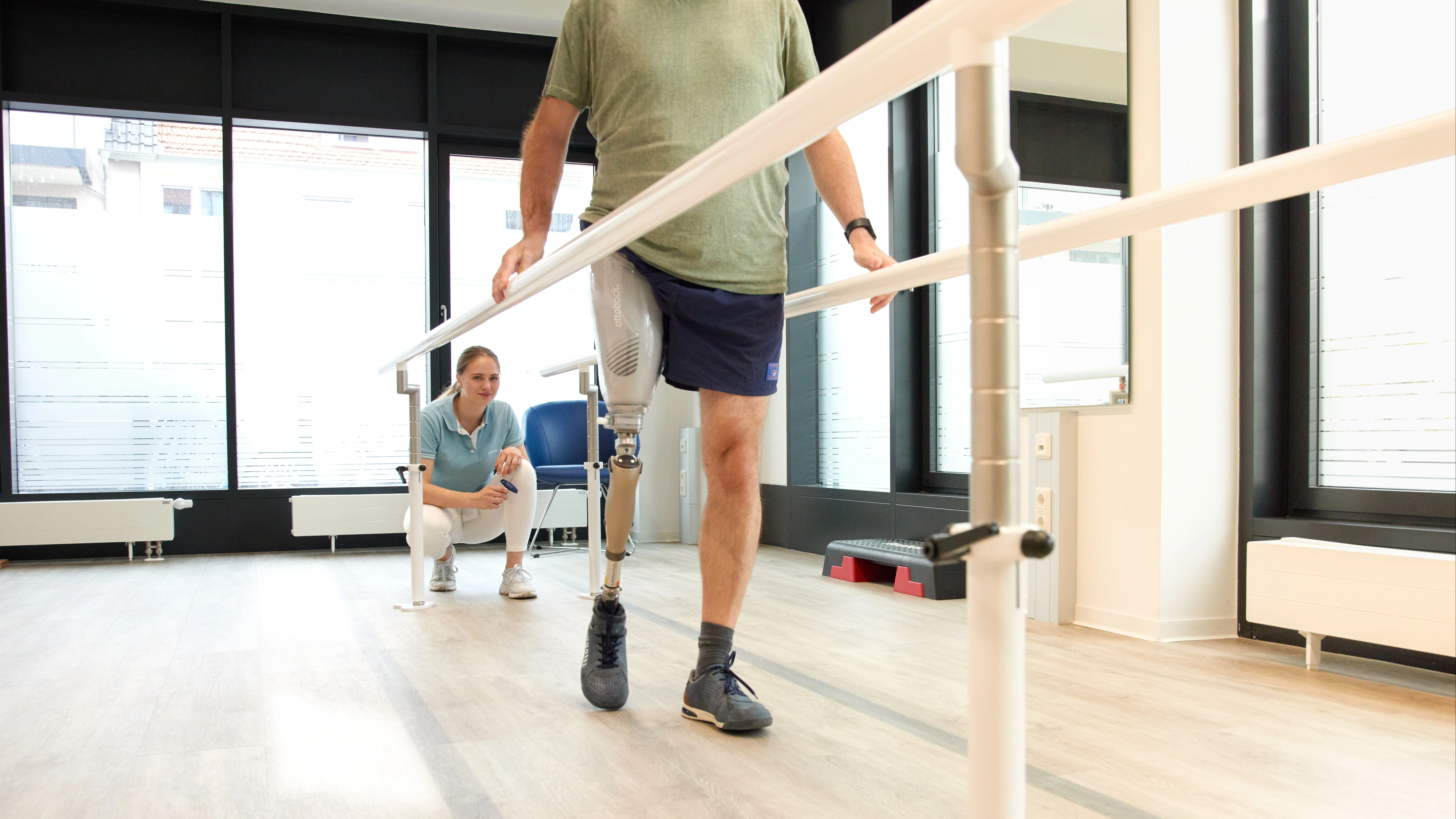 Man with above knee leg prosthesis walking between parallel bars during gait training, physiotherapist supervising in a rehabilitation clinic