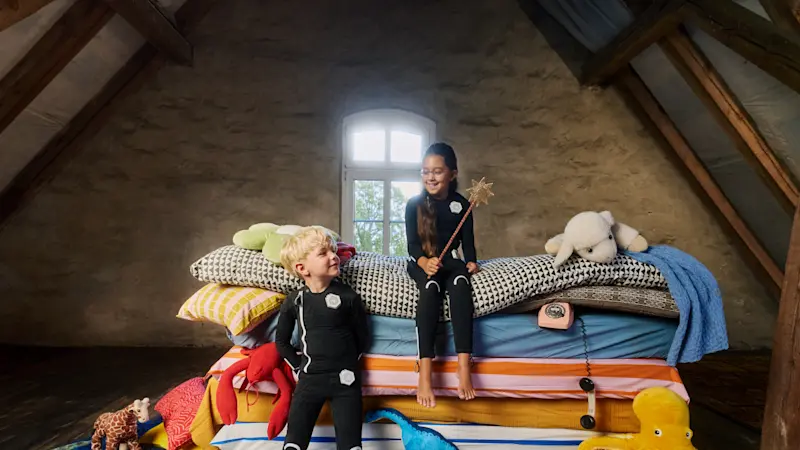 Two children wearing Exopulse neuromodulation suits sitting on a colorful bed surrounded by toys during pediatric therapy session