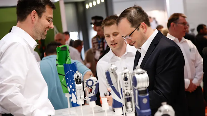 In einem mit Menschen gefüllten Raum stehen drei Männer an einem weißen Tisch und begutachten die darauf ausgestellten Innovationen von Ottobock.
