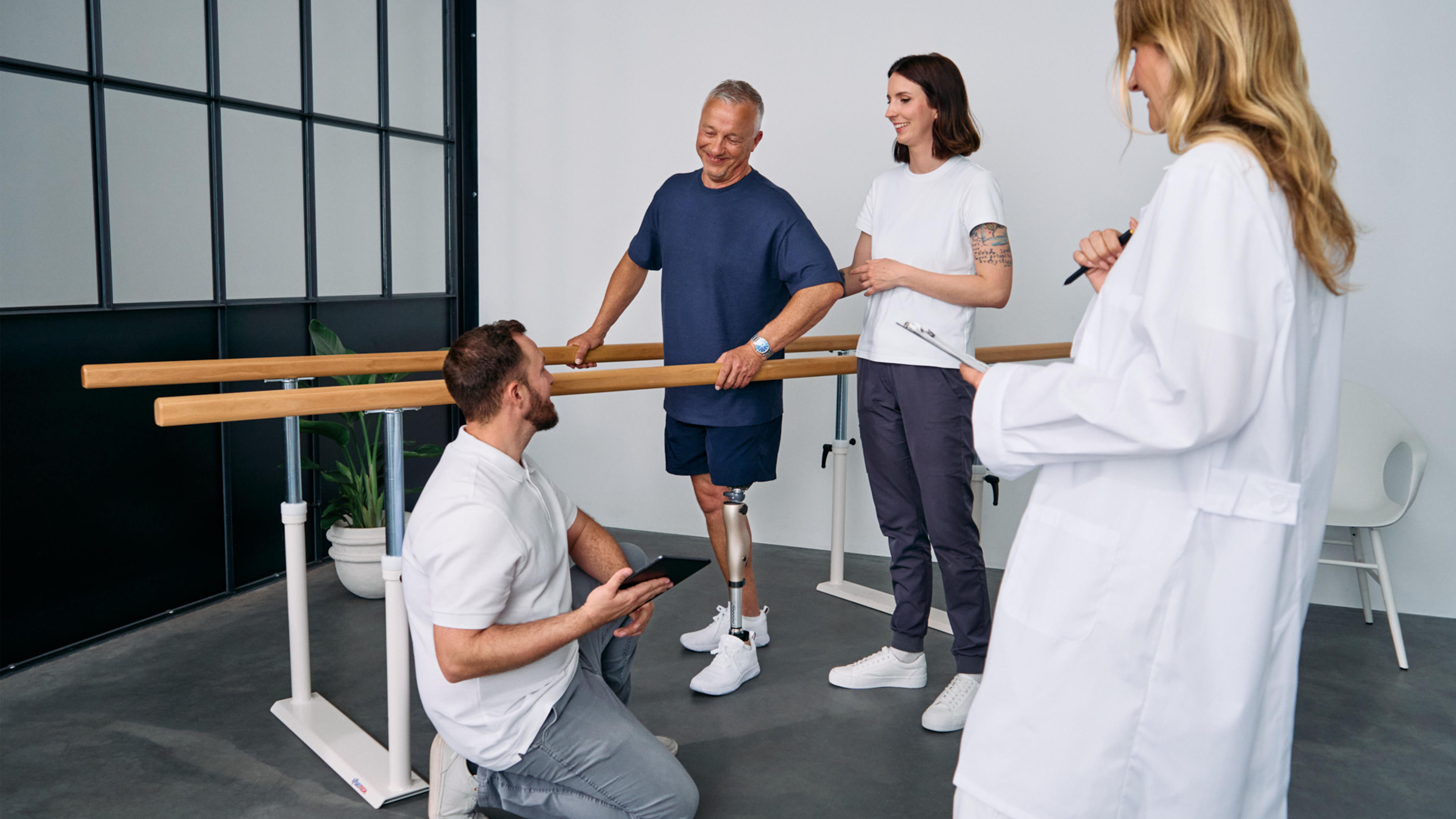 Patient with below knee leg prosthesis standing at parallel bars, rehabilitation team reviewing progress during prosthetic gait training session