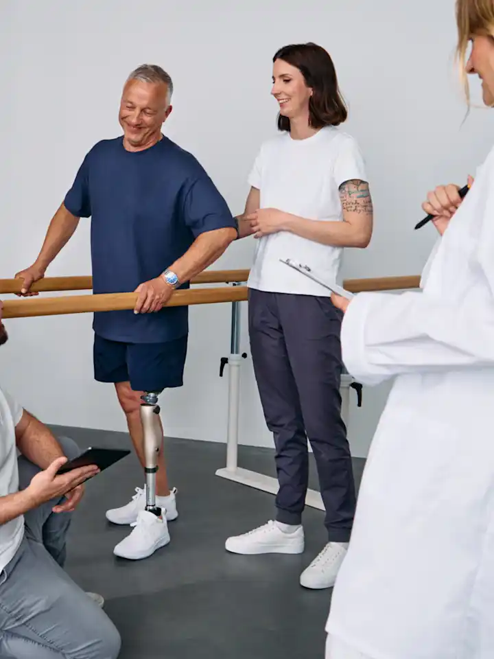 Patient with below knee leg prosthesis standing at parallel bars, rehabilitation team reviewing progress during prosthetic gait training session