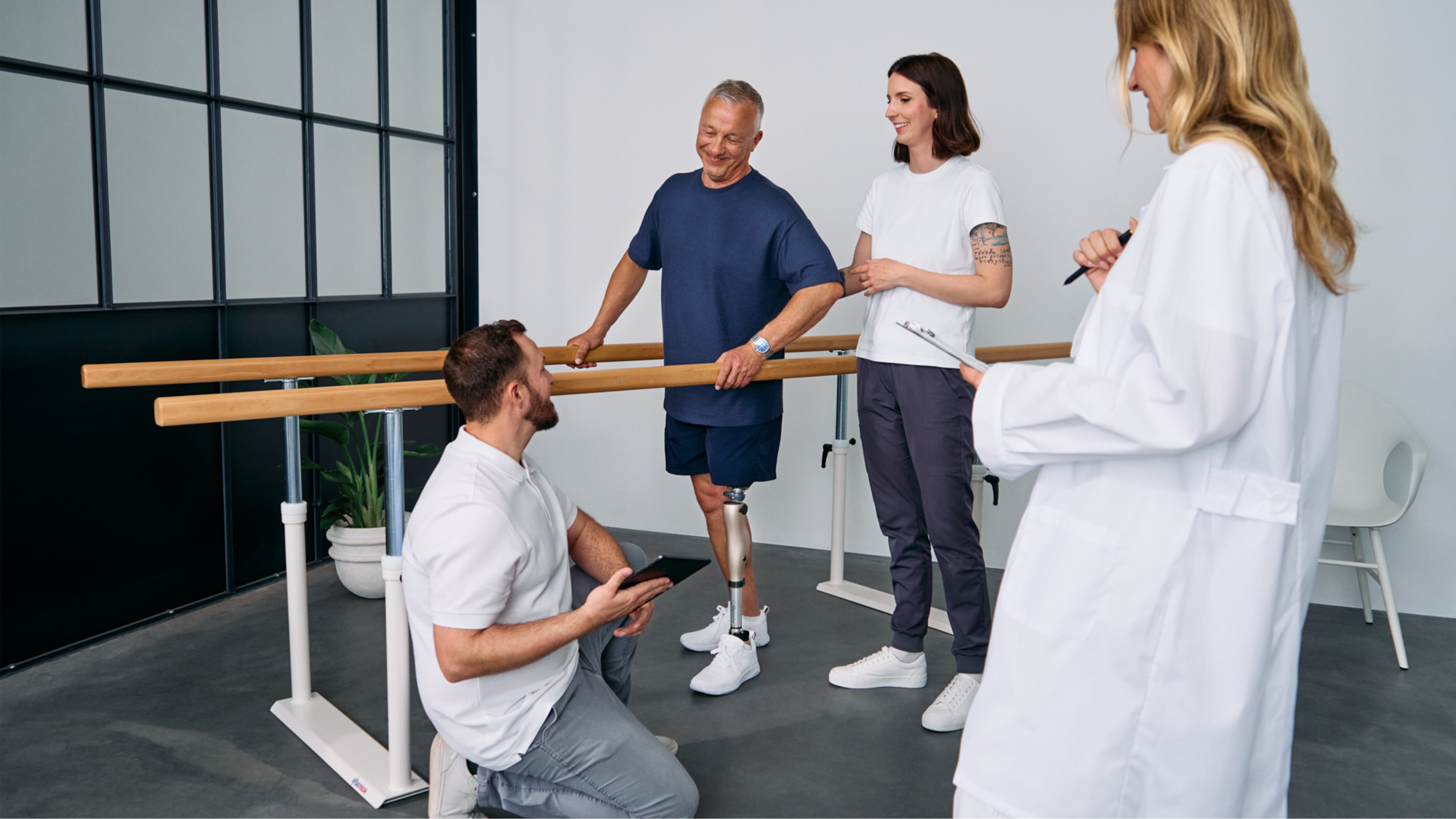 Patient with below knee leg prosthesis standing at parallel bars, rehabilitation team reviewing progress during prosthetic gait training session