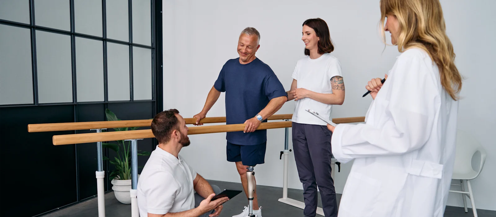 Patient with below knee leg prosthesis standing at parallel bars, rehabilitation team reviewing progress during prosthetic gait training session