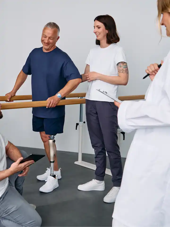 Patient with below knee leg prosthesis standing at parallel bars, rehabilitation team reviewing progress during prosthetic gait training session