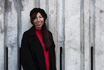 a photograph of Esther Jonson, a white woman with dark hair, smiling in front of a poured concrete wall. 