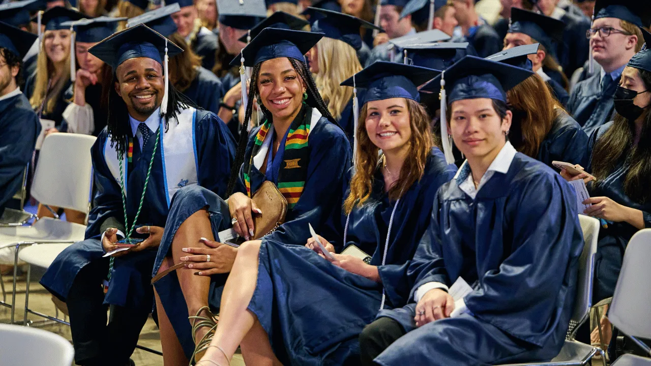 Four Penn State Smeal graduates sit next to each other during a commencement ceremony in their blue caps and gowns.