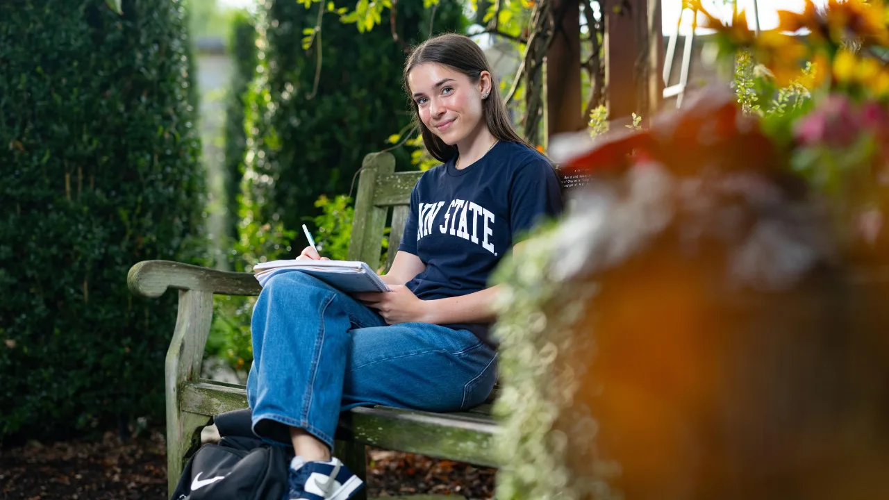 A student writing outside on a bench smiles
