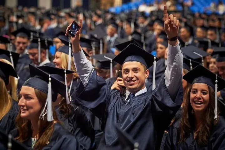 Graduating student in regalia raises arms and smiles in a crowd of graduates