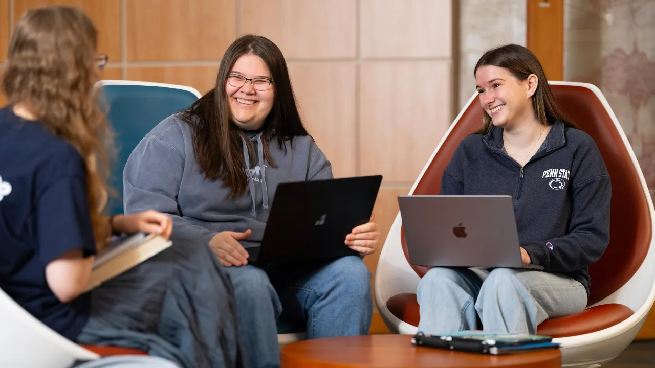 Three students sit in the lobby and smile over laptops