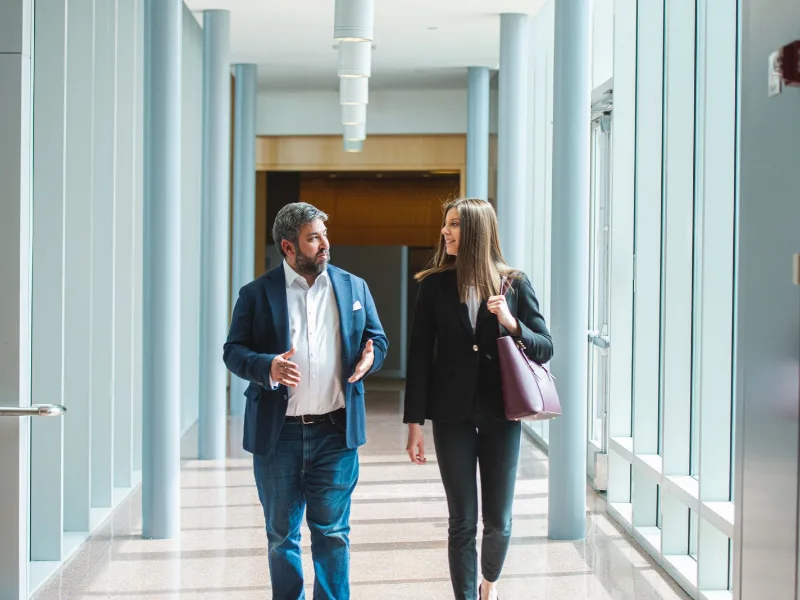 Two people in professional attire talk as they walk down a hallway