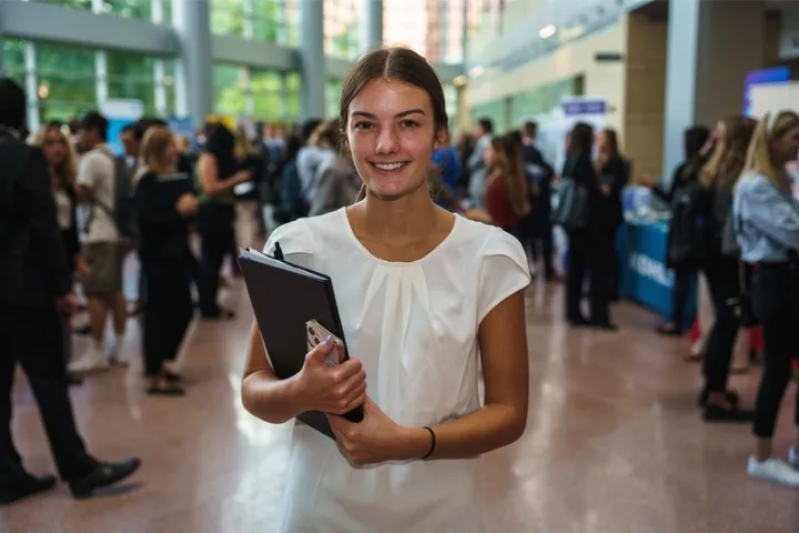 Student in professional attire smiles with career fair crowd in background